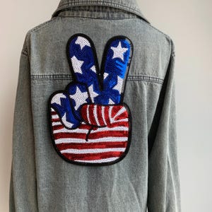 Può includere: Una giacca di jeans con una grande toppa a forma di segno della pace cucita sul retro. La toppa è realizzata con paillettes rosse, bianche e blu e presenta un segno della pace con il design della bandiera americana.