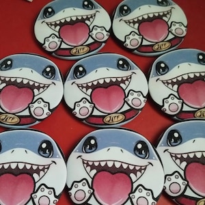 May include: A group of circular buttons with a cartoon shark design. Each button shows a smiling shark with a blue head, white teeth, and a pink heart-shaped tongue. The buttons have a red and white border with the name "JEFF".