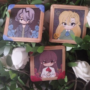 Op de afbeelding: Drie vierkante pins met anime-stijl karakterillustraties. Eén toont een grijsgekleurd personage met blauwe rozen, een andere een blond personage, en de derde een donkerharig personage dat een rode roos vasthoudt, allemaal in goudkleurige kaders.