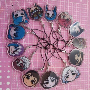May include: A collection of 12 round, clear acrylic charms featuring cartoon characters with different expressions and hairstyles. Each charm has a black string attached to a silver metal ring.