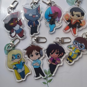 May include: A selection of acrylic keyrings showcasing cartoon characters. The keyrings have silver clasps and feature diverse figures with distinct designs and colour schemes. The characters include a devil, a cat in a suit, and a superhero.