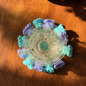May include: A clear glass teacup with a floral design sits on a handmade crochet coaster. The coaster features a scalloped edge in shades of blue and purple. The teacup is filled with a clear liquid.