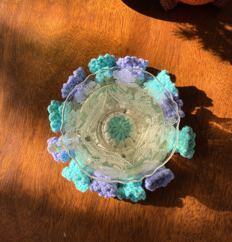 May include: A clear glass teacup with a floral design sits on a handmade crochet coaster. The coaster features a scalloped edge in shades of blue and purple. The teacup is filled with a clear liquid.