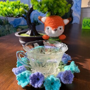 May include: A clear glass teacup with a floral design sits on a crocheted coaster with blue and purple flower accents. A small, orange and white fox figurine is in the background, near a bonsai tree.