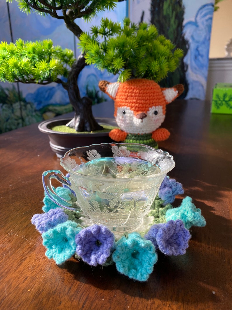 May include: A clear glass teacup with a floral design sits on a crocheted coaster with blue and purple flower accents. A small, orange and white fox figurine is in the background, near a bonsai tree.