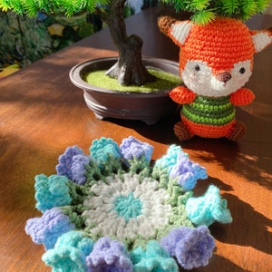 May include: A handmade crochet coaster with a floral design in shades of blue, green, and white. A small, orange and white crocheted fox toy is next to the coaster. A small bonsai tree is in the background.