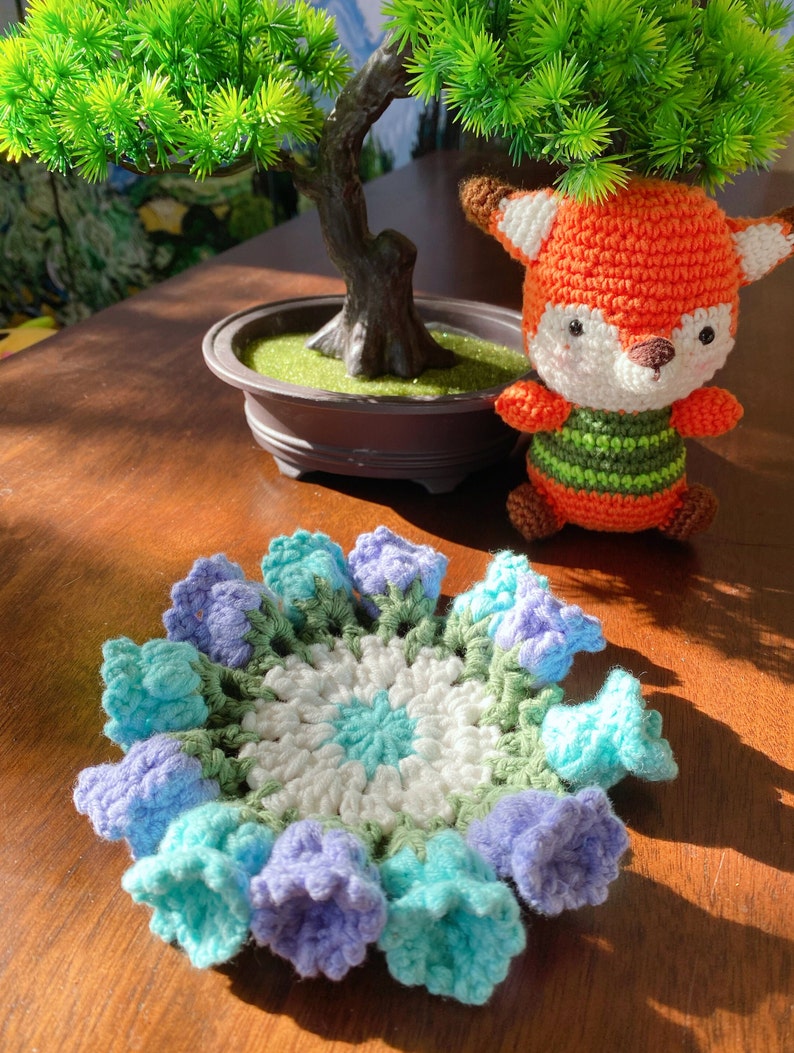 May include: A handmade crochet coaster with a floral design in shades of blue, green, and white. A small, orange and white crocheted fox toy is next to the coaster. A small bonsai tree is in the background.