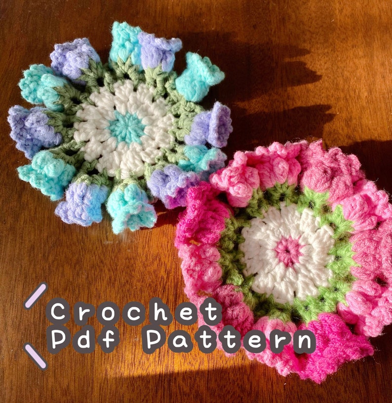 May include: Two handmade crochet flower coasters. One coaster features a white center with light blue, green, and purple flower petals. The other coaster has a white center with pink and green flower petals. The text "Crochet Pdf Pattern" is also visible.