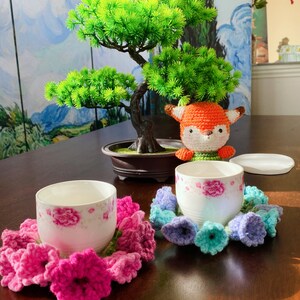 May include: Two white teacups with pink floral designs sit on a dark wooden table, each surrounded by a crocheted flower coaster. A crocheted fox and a small bonsai tree are also on the table. The coasters are in shades of pink, blue, and green.