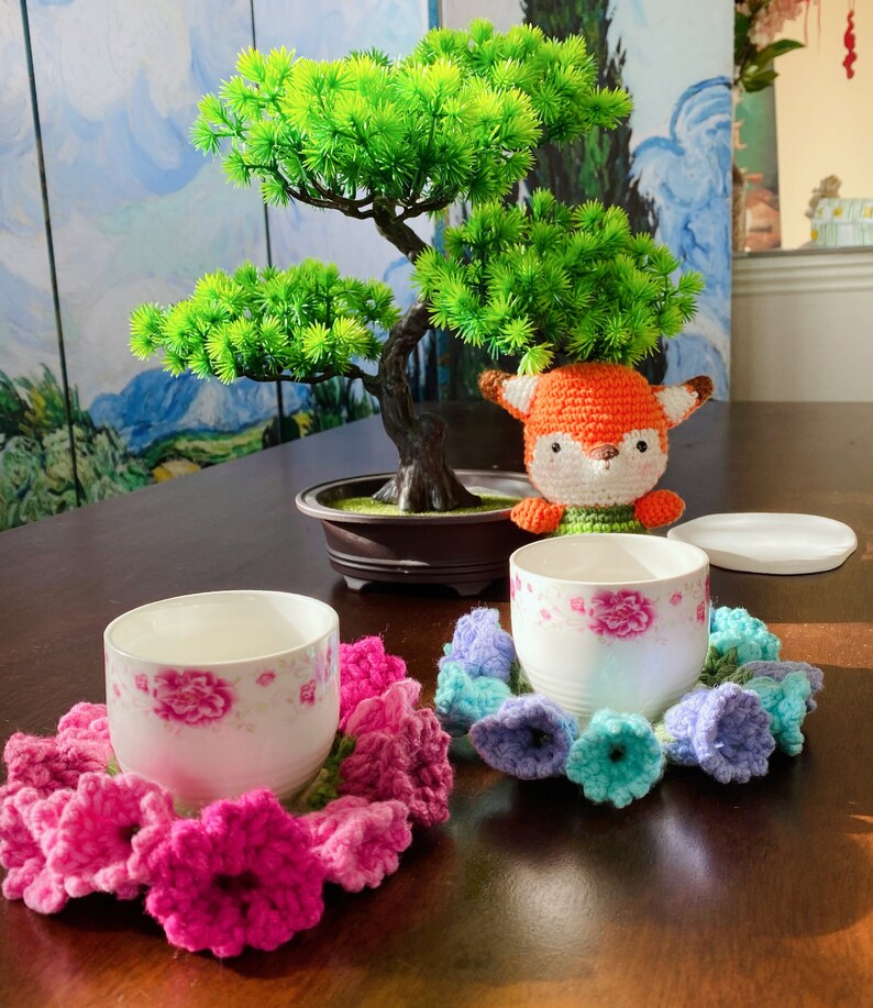 May include: Two white teacups with pink floral designs sit on a dark wooden table, each surrounded by a crocheted flower coaster. A crocheted fox and a small bonsai tree are also on the table. The coasters are in shades of pink, blue, and green.