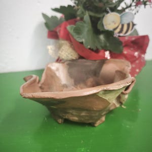 Puede incluir: Una maceta de cerámica marrón hecha a mano con un borde ondulado único. Contiene una pequeña planta con flores rojas y hojas verdes. Se adjunta una pegatina decorativa de abeja. La maceta se encuentra sobre una superficie verde.