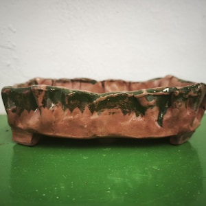 Puede incluir: Una maceta rectangular de cerámica para bonsái con una textura irregular hecha a mano. La base es de color arcilla terracota natural, mientras que el borde superior presenta un esmalte verde oscuro grueso que gotea por los lados.