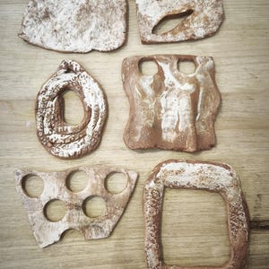Puede incluir: Una colección de seis formas de arcilla de terracota moldeadas a mano con bordes irregulares y superficies blancas texturizadas. Las piezas incluyen varias formas geométricas, algunas con recortes circulares, bucles y centros abiertos.