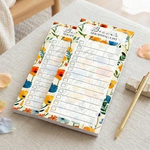 Puede incluir: Dos blocs de notas de lista de compras florales con el texto "Becca's SHOPPING LIST". Los blocs presentan un fondo crema con diseños florales naranjas, amarillos y azules. También hay un bolígrafo y una goma de borrar dorados.