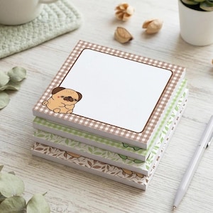 May include: A stack of square notepads with a brown and white gingham border and a cute pug illustration. The notepads have a white center for writing and are stacked on top of each other. A silver pen rests on the side.