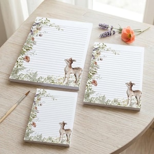 Pode incluir: Três blocos de notas pautados de vários tamanhos, cada um com uma ilustração em aquarela de um veado e uma borda floral. Os blocos são brancos com linhas cinzentas e um design botânico em tons de verde, castanho e pêssego.