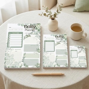 Peut inclure: Ensemble de trois blocs-notes planificateurs quotidiens de différentes tailles, avec un motif de feuilles d'eucalyptus vert et blanc. Chaque bloc-notes comprend des sections pour les listes de tâches, les rendez-vous et les notes. Un stylo en bois et une tasse de thé sont également présents.