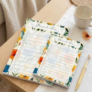 Puede incluir: Dos blocs de notas con motivos florales y el texto "Becca's notes" están sobre una mesa de madera. Los blocs tienen un diseño de lista de verificación y están adornados con flores de colores en tonos naranja, amarillo y azul. También se ve un bolígrafo dorado y una taza de café.