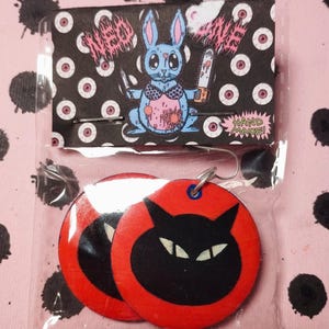 Può includere: Un paio di orecchini in legno rosso e nero con un design a forma di gatto nero. Gli orecchini sono fatti a mano e vengono forniti in una busta di plastica trasparente con una carta che dice "HAND MADE!" e raffigura un coniglio dei cartoni animati con una motosega e un coltello.