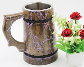 Könnte beinhalten: Ein hölzerner Bierkrug mit Edelstahl-Innenraum und dekorativem Griff. Der Krug hat eine braune Oberfläche mit lila Akzenten und schwarzen Seildetails. Ein kleiner weißer Topf mit roten Rosen und weißen Blumen steht daneben.