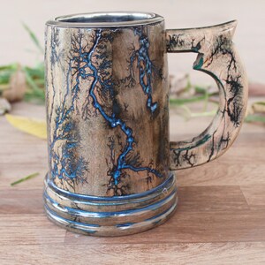 May include: A cylindrical wooden mug featuring a natural light brown wood grain finish with intricate black fractal burn patterns and vibrant blue resin inlay. The mug includes a sturdy curved handle and a smooth metallic interior lining.