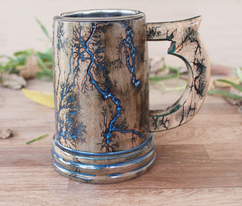 May include: A cylindrical wooden mug featuring a natural light brown wood grain finish with intricate black fractal burn patterns and vibrant blue resin inlay. The mug includes a sturdy curved handle and a smooth metallic interior lining.