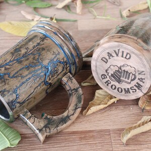 May include: Two wooden beer mugs featuring Lichtenberg wood burning patterns. One mug displays blue resin-filled lightning-like fractures, while the other base is engraved with the text DAVID GROOMSMAN and an illustration of two beer mugs.