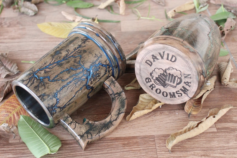 May include: Two wooden beer mugs featuring Lichtenberg wood burning patterns. One mug displays blue resin-filled lightning-like fractures, while the other base is engraved with the text DAVID GROOMSMAN and an illustration of two beer mugs.