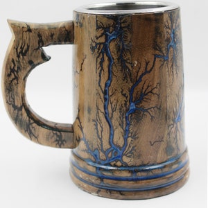 May include: A wooden beer mug featuring a natural brown finish with intricate, branching fractal patterns burned into the surface and filled with a vibrant blue resin. The mug includes a sturdy handle and a smooth, metallic silver-colored interior rim.