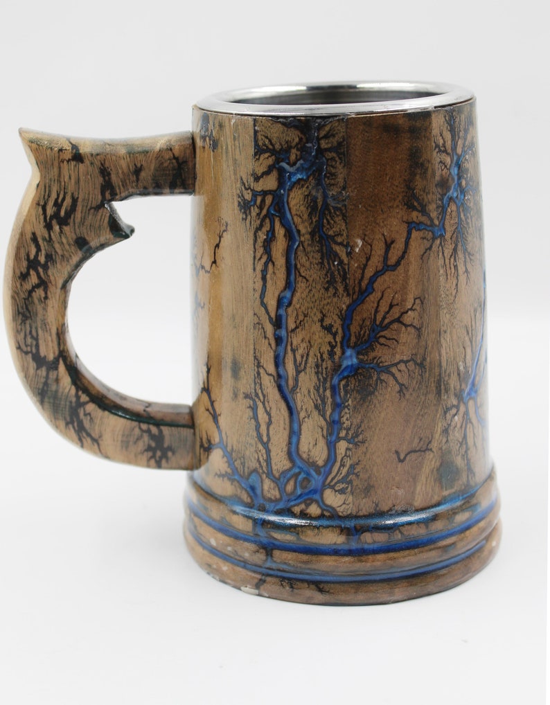 May include: A wooden beer mug featuring a natural brown finish with intricate, branching fractal patterns burned into the surface and filled with a vibrant blue resin. The mug includes a sturdy handle and a smooth, metallic silver-colored interior rim.