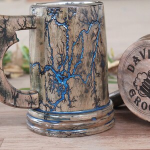 May include: A wooden beer mug featuring intricate black fractal burn patterns and a vibrant blue resin inlay. The base of the mug is engraved with the text DAVID GROOMSMAN and an illustration of two beer mugs clinking together.