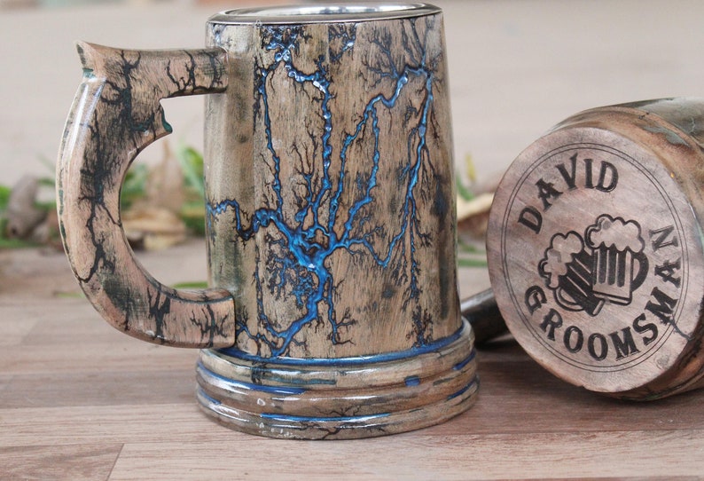 May include: A wooden beer mug featuring intricate black fractal burn patterns and a vibrant blue resin inlay. The base of the mug is engraved with the text DAVID GROOMSMAN and an illustration of two beer mugs clinking together.