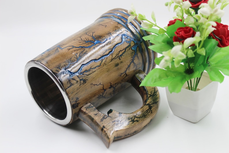 May include: A cylindrical mug with a polished silver-toned metal interior and a wooden exterior featuring dark, branching fractal patterns filled with a vibrant blue resin inlay. The mug has a sturdy wooden handle and a smooth, glossy finish.