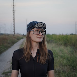 May include: A person wearing a black cycling jersey, large black framed glasses, and a navy blue cycling cap with the text "NEED COFFEE NOW". The background features a dirt path and grassy field.