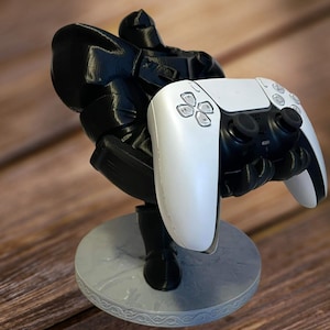 May include: A black 3D printed figure holding a white PlayStation 5 controller. The figure is standing on a round base.