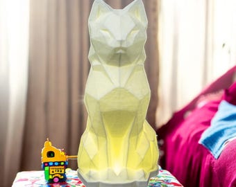 May include: A white geometric cat lamp with a light yellow glow. The lamp is sitting on a colorful surface.