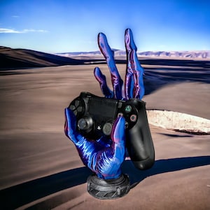 May include: A blue and purple hand-shaped gaming controller holder. The holder is designed to look like a hand making a peace sign, holding a black gaming controller. The base of the holder is grey.