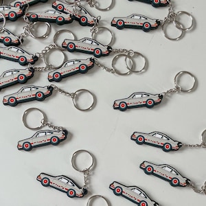 Puede incluir: Una colección de llaveros de coche blancos con detalles rojos y negros. Los llaveros presentan un diseño de coche clásico y están unidos a llaveros de plata.