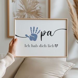 Puede incluir: Un cuadro enmarcado con la palabra "pa" en cursiva, una huella de mano azul y la frase "Ich hab dich lieb" en negro. El cuadro se muestra sobre un fondo de tonos neutros.