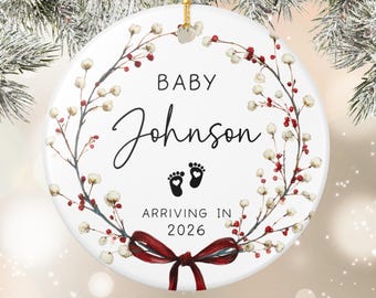 May include: A white ceramic Christmas ornament with a floral wreath design. The ornament features the text "BABY Johnson ARRIVING IN 2026" and two black baby footprints. A red ribbon is at the bottom.
