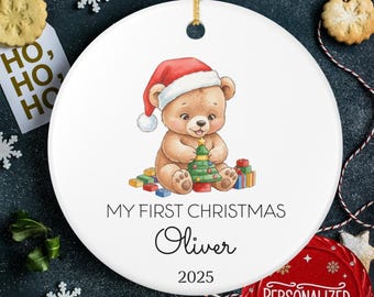 May include: White ceramic Christmas ornament featuring a brown teddy bear wearing a Santa hat, holding a Christmas tree, and surrounded by colorful presents. The text reads "MY FIRST CHRISTMAS" and "Oliver 2025".