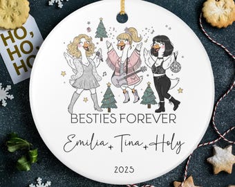 May include: White ceramic Christmas ornament with a gold string. The ornament features a cartoon illustration of three geese dressed in festive attire, with the text "BESTIES FOREVER" and names. The year "2025" is also printed on the ornament.