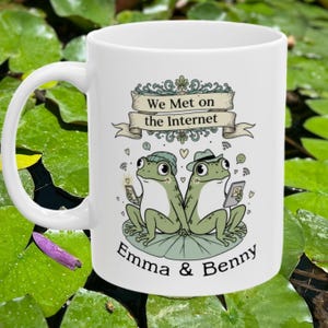 May include: White ceramic mug featuring a whimsical illustration of two green frogs sitting on a lily pad. The mug has the text "We Met on the Internet" and the names "Emma & Benny". The frogs are holding phones and tablets.
