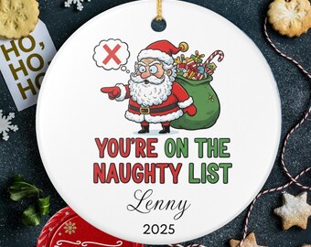 May include: A white ceramic Christmas ornament featuring Santa Claus pointing with a thought bubble with an X. Text reads "YOU'RE ON THE NAUGHTY LIST" with the name "Lenny" and the year "2025". The ornament has a gold string for hanging.