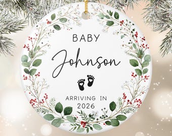 May include: A white ceramic ornament with a gold string for hanging. The ornament features the text "BABY Johnson" in black script, baby footprints, and the text "ARRIVING IN 2026". It is decorated with a wreath of green leaves and red berries.