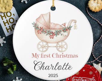 May include: White ceramic Christmas ornament featuring a baby carriage illustration in soft pink and beige tones. Text reads "My first Christmas Charlotte 2025". Includes a gold hanging cord.