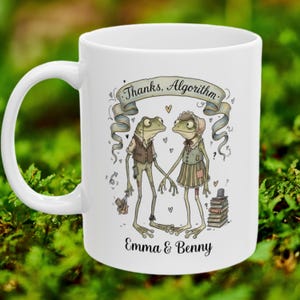 May include: White ceramic mug featuring a whimsical illustration of two frog characters holding hands. The mug has the text "Thanks, Algorithm" above the frogs and "Emma & Benny" below. The frogs are wearing clothes.