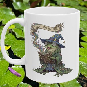 May include: White ceramic mug featuring a whimsical illustration of a frog wizard reading a spell book. The frog wears a blue hat with stars and a green robe. The banner above the frog reads "I didn't read the spell."