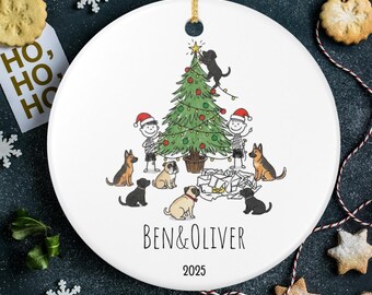 May include: White ceramic Christmas ornament with a festive illustration. The design features a Christmas tree decorated with lights, two children, and several dogs. The names "BEN & OLIVER" and the year "2025" are printed below the illustration. A gold string is attached for hanging.