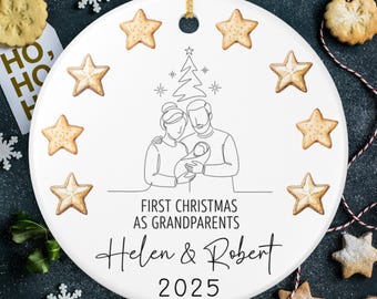 May include: White ceramic Christmas ornament with a gold string for hanging. The ornament features a line drawing of a family with a baby, surrounded by star-shaped cookies. Text reads "First Christmas as Grandparents, Helen & Robert, 2025."